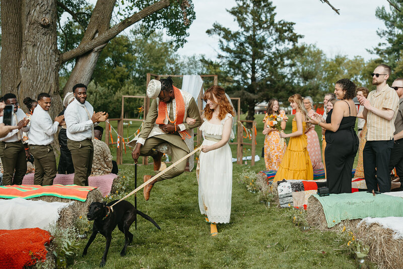 Vibrant outdoor wedding featuring a couple and their dog walking down the aisle while the groom jumps and clicks his heels, surrounded by colorful decor.