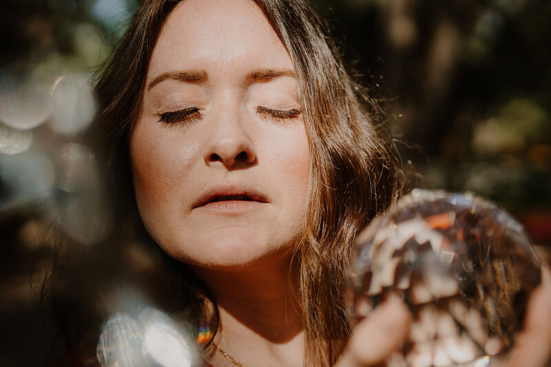 The Intuitive Momma stands in the forest and closes eyes and sun reflects light on a crystal ball in her hands