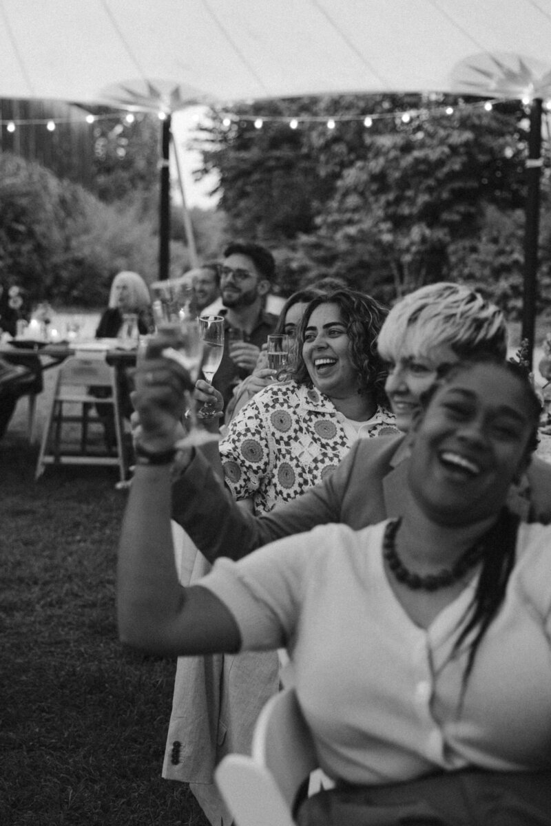 black and white candid photo of guests holding up drinkins at wedding reception during speeches