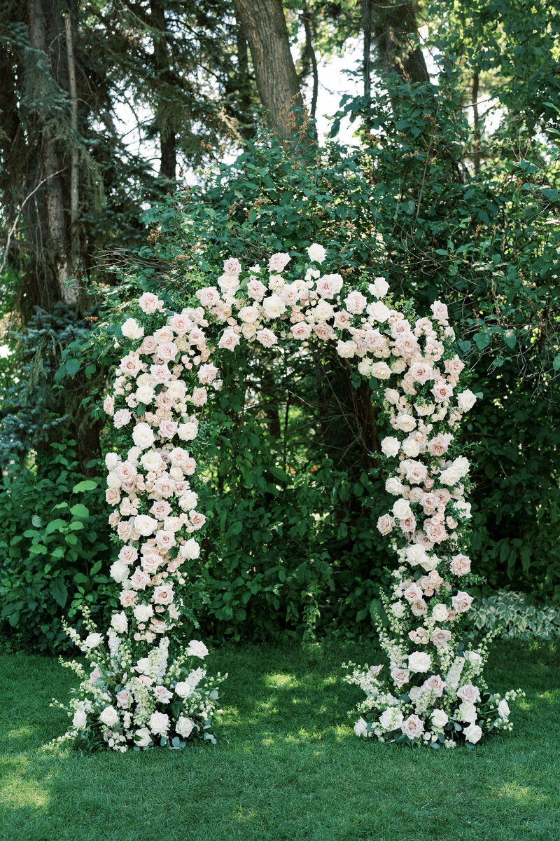 A flower arch made of numerous pink and white roses set against a backdrop of lush green trees and plants, beautifully crafted by a full service wedding planner. Two stunning flower arrangements stand symmetrically at the base of the arch.