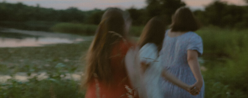 Three friends are holding hands as they run next to a pond with lily pads and a beautiful sunset