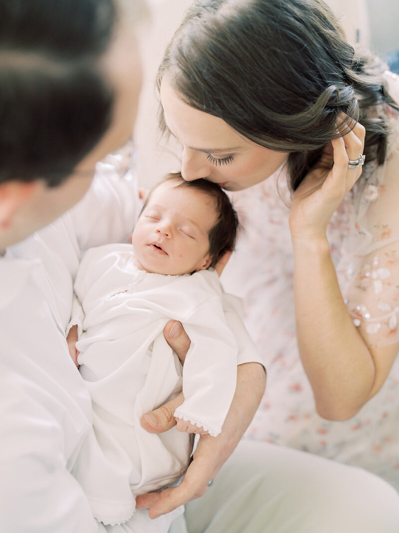 Mother with brown hair leans down to kiss baby girl in white onesie held by her husband during her newborn session by Marie Elizabeth Photography.