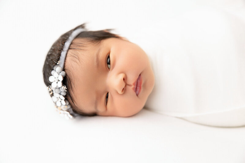 Baby girl posed for newborn photo session wrapped in white and wearing flowered headband. 