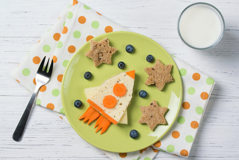 Aerial photo of a healthy lunch for a child including a sandwich made to look like a rocket ship and blueberries to look like stars served with millk