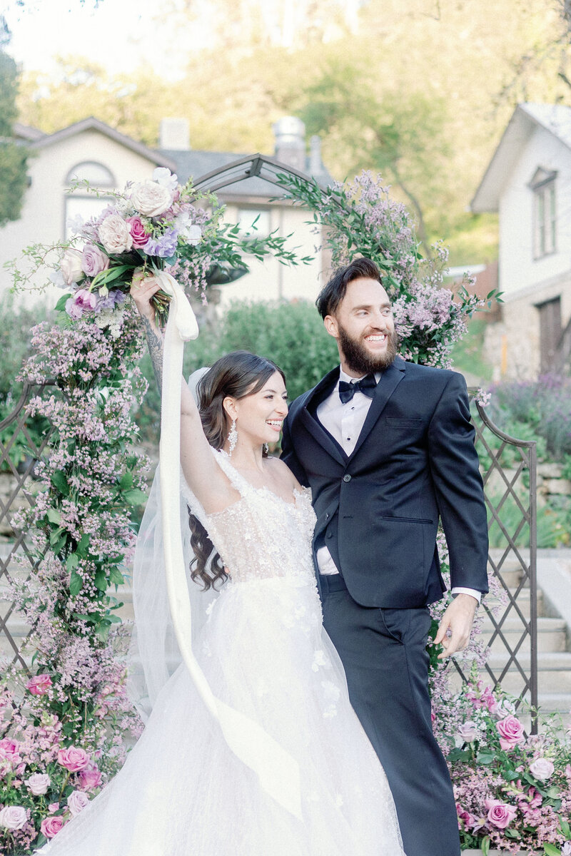 bay area photographers captures bride adn groom celebrating after their ceremony with bride holding her bouquet in the air as her groom holds her and smiles with a wedding arch covered in florals