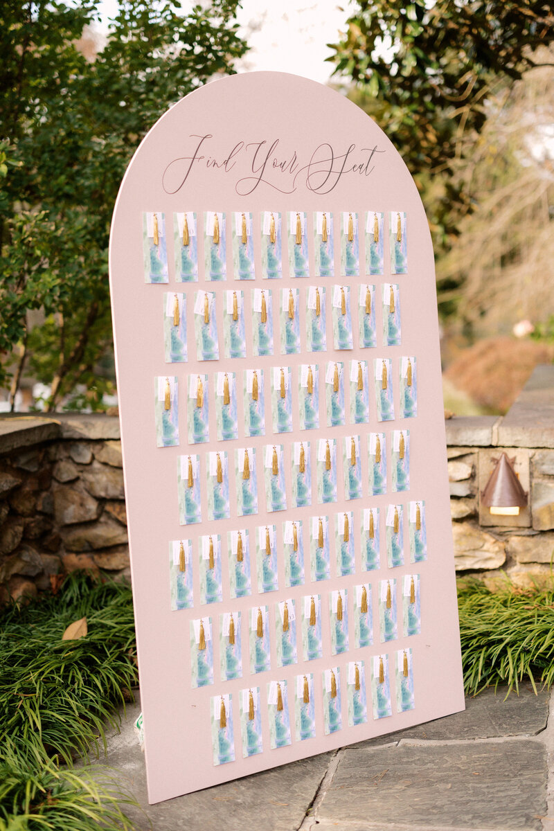 November wedding at Graylyn Estate, designed and planned by Charming Carolina — personalized bookmark escort display featuring watercolor artwork by the groom’s brother, gold tassels, table numbers, and a thank you note from the couple. Each bookmark hung on a blush board as a creative guest seating display.