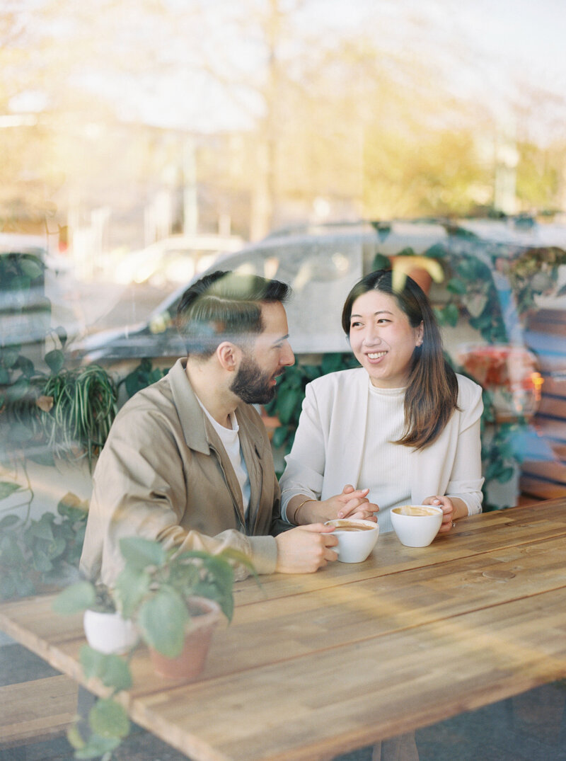 seattle engagement photographer captures  couple smiling at each other while drinking coffee, photo taken through a window