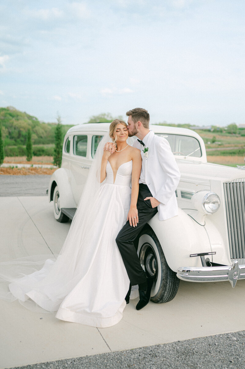 Bride and Groom lean on vintage get away car on their wedding day.