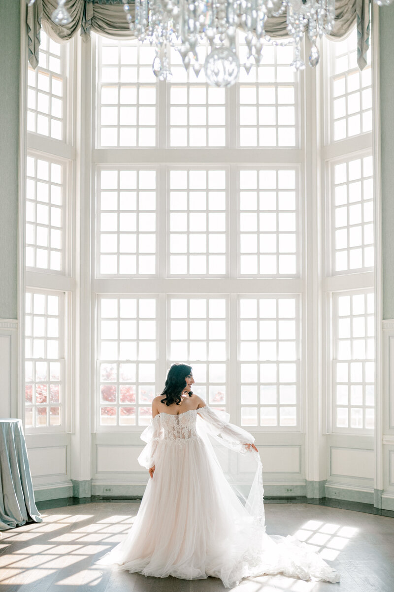 Bride in a flowing wedding gown standing by large windows, captured by a Virginia Wedding Photographer in a beautifully lit Estate at River Run in Maidens, Virginia