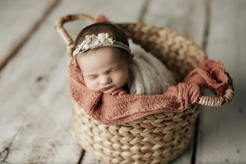 Baby girl sleeping in woven basket in for newborn portraits in top Exeter, Ontario photo studio. Basket is draped  in an apricot muslin swaddle and baby is wrapped in a cream knit swaddle  with matching headband with petite cream flowers. Baby