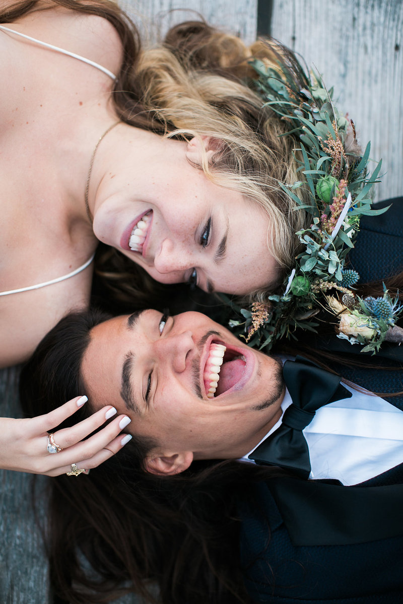 bride and groom laughing on wedding day while laying down on a dock