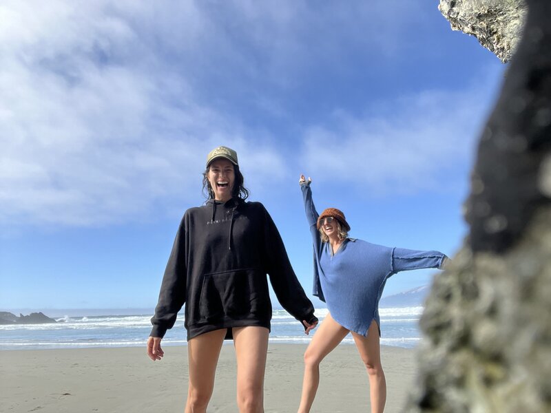 "Two women enjoying a sunny day at the beach, smiling and posing playfully, with a blue sky, ocean waves, and sandy shore in the background."