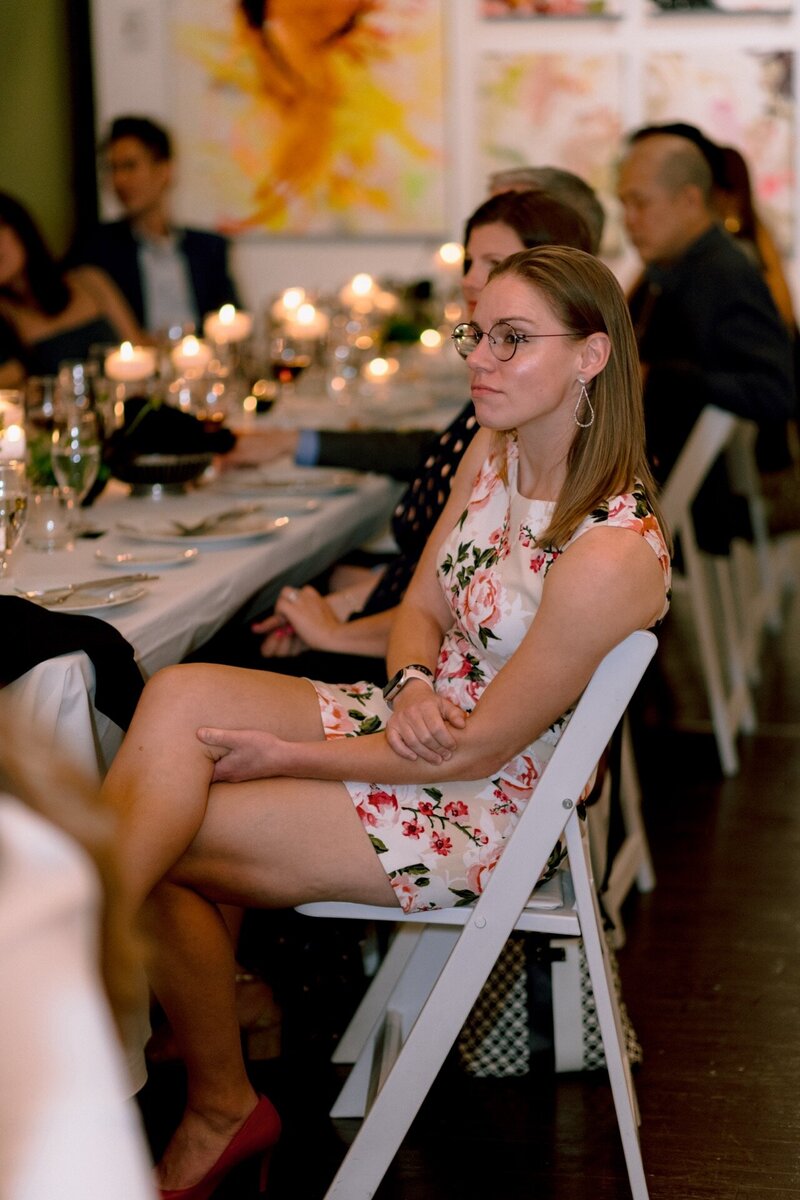 A woman in a floral dress sitting at a banquet table with other guests in the background.