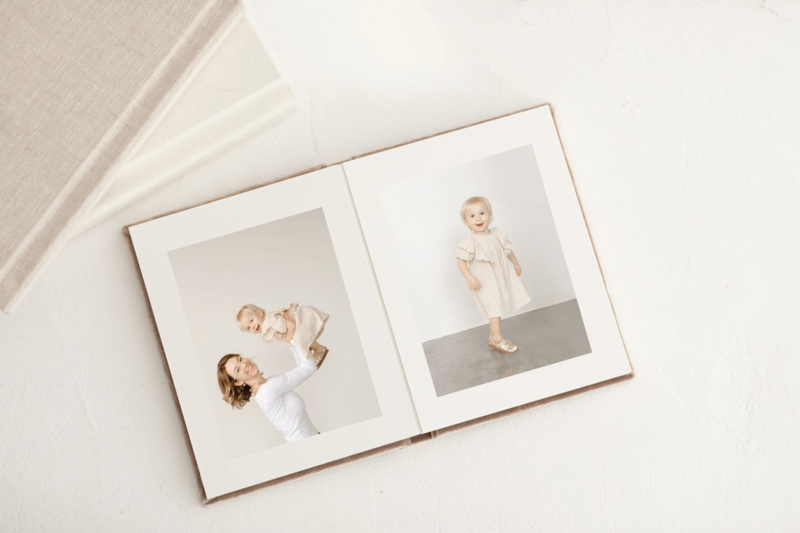 Family of three sitting in a field of golden grasses with their small little dog as the sunsets. This picture is within a photo album and there are some other linen photo albums stacked up next to the one laying open.