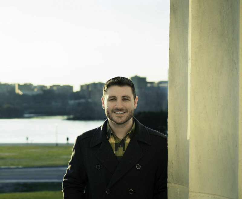 A man in a dark peacoat and a yellow plaid shirt smiles warmly, standing by a large column with a scenic river and cityscape in the soft glow of sunset in the background. His casual yet stylish attire and the serene outdoor setting create a pleasant and inviting atmosphere.