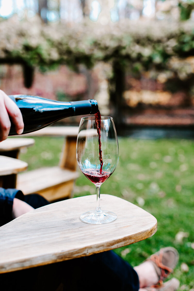 Close up image of red wine pouring into a wine glass sitting on the arm of a wooden chair | Sara Coffin Photo in NC