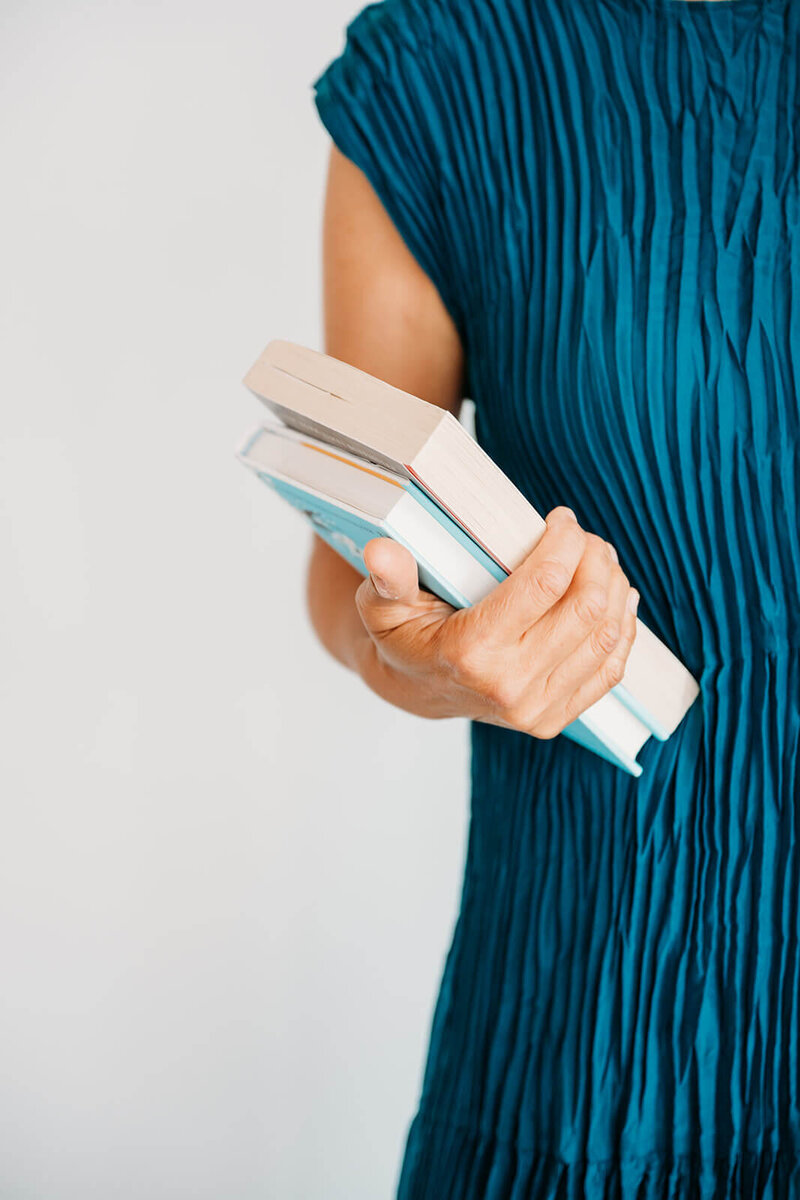 Close-up of a person’s hand holding a stack of books against a teal pleated dress.