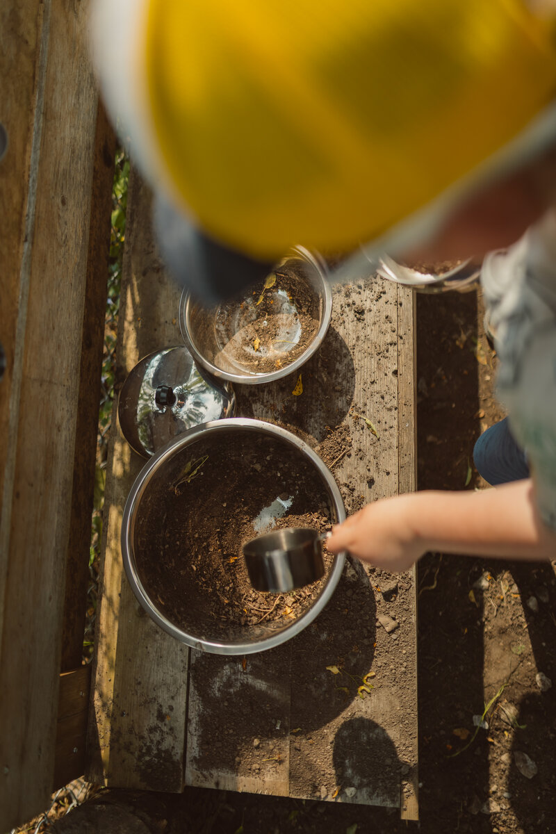 photo from the perspective above a young boy playing in a mud kitchen scooping dirt with a measuring cup into a large metal basin
