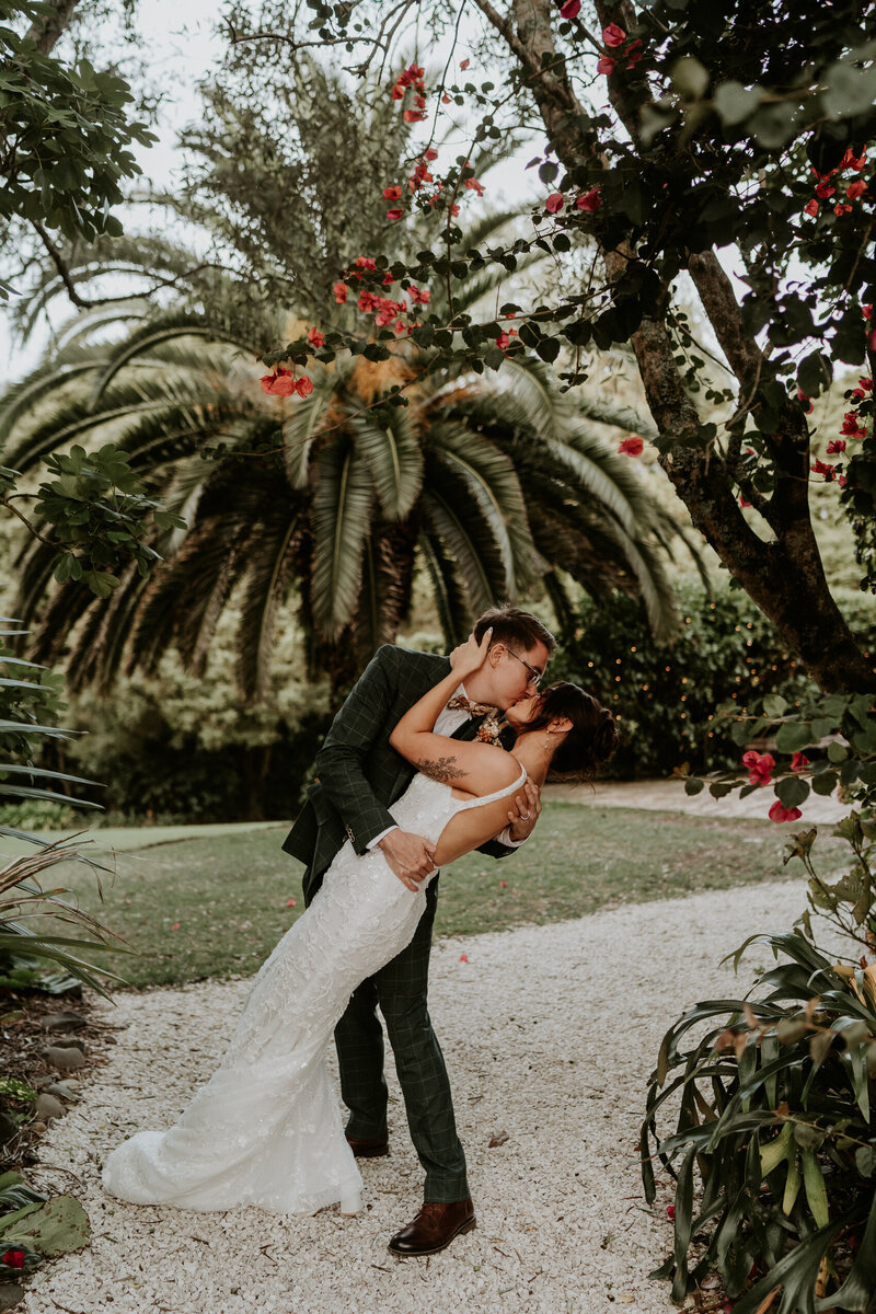 Bride and groom share a romantic kiss under palm trees and florals in a tropical garden setting at their elegant Auckland wedding