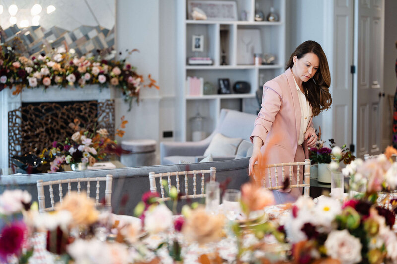 wedding planner emma westacott straightens chairs at a dining table decorated with peach and burgundy flowers