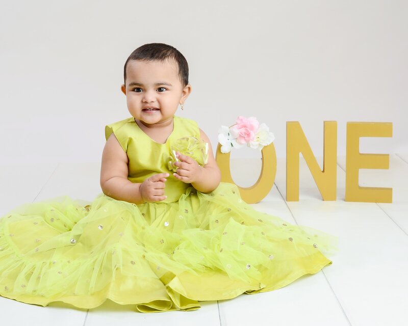 A baby girl in a beautiful green dress, holding a small flower prop, sitting next to golden "ONE" letters during her first birthday photoshoot.