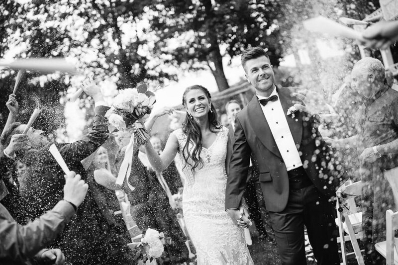 Deux mariés sous une pluie de confettis lors de leur mariage à l'extérieur au Manoir Hovey, image en noir et blanc. 
