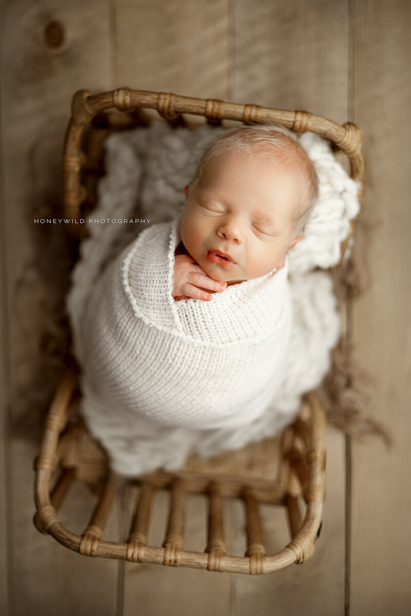 Newborn baby swaddled in a white knit wrap, peacefully sleeping in a wicker basket on a rustic wood floor. Photographed by Honeywild Photography, a luxury newborn photographer in Grand Rapids, MI, specializing in timeless and elegant portraits
