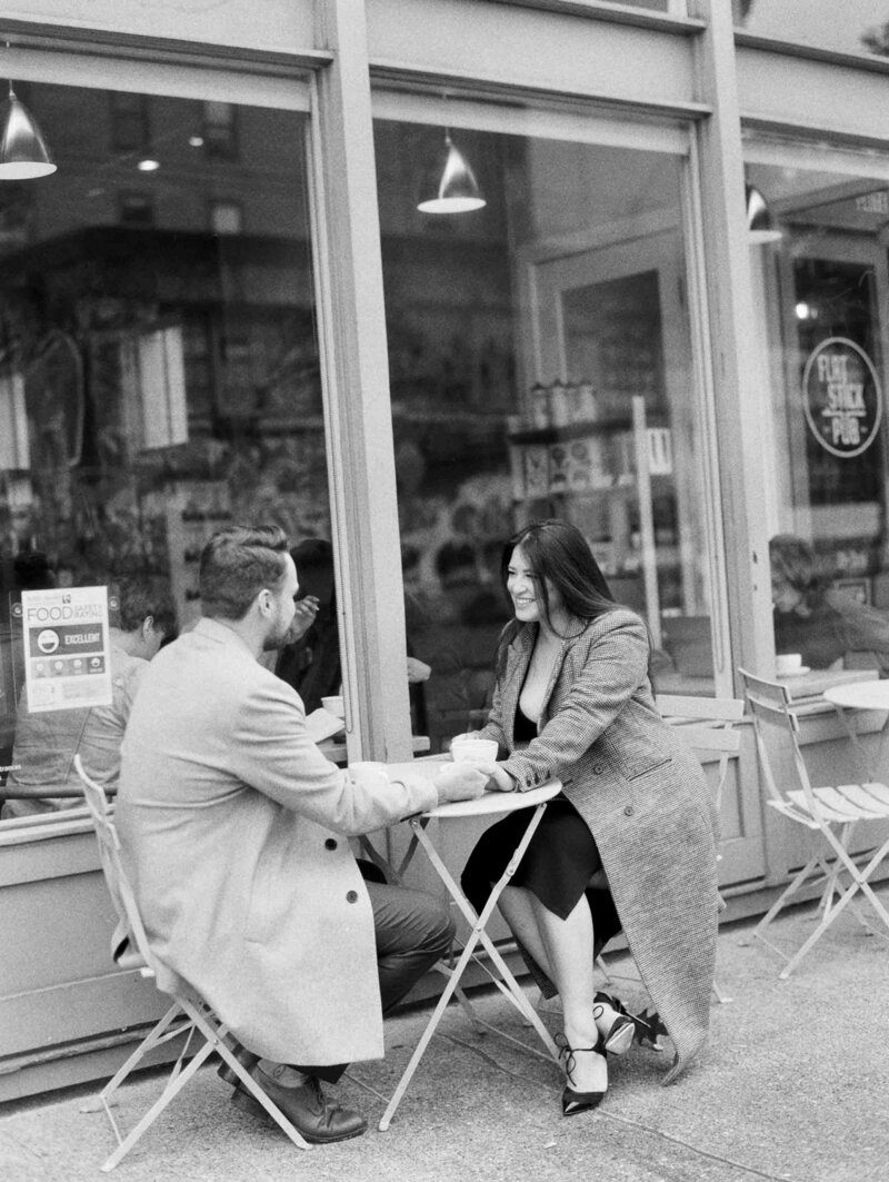 Seattle engagement photographers capture black and white image of happy couple sitting at cafe table out front of a coffee shop in seattle on black and white film