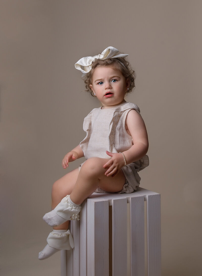 A little toddler girl is sitting atop of a white wooden crate for a studio portrait in brooklyn, ny.  She is wearing a beige romper with a big beige bow in her curly hair. She is looking intently at the camera. Captured by top Brooklyn, NY family photographer Chaya Bornstein Photography.