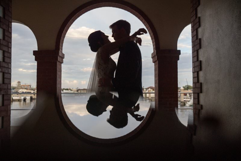 Silhouetted wedding couple embracing through an oval archway at the White Room in St. Augustine, with their reflection visible in calm water below and marina boats in the background under a cloudy sky