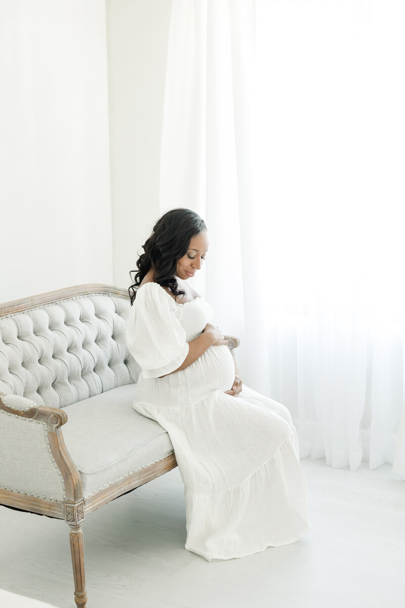 Pregnant woman wears white flowing dress and sits on a settee by a window while embracing her belly during maternity portrait session