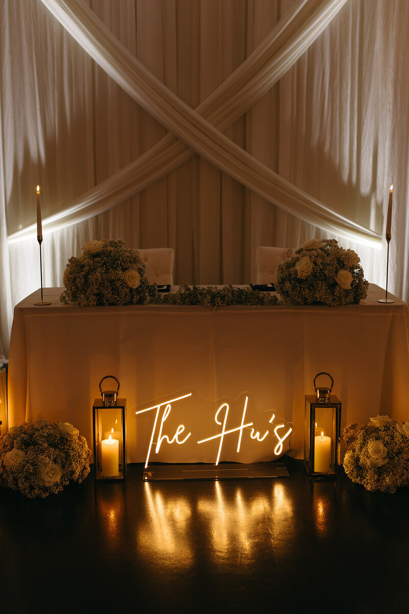 A beautifully decorated sweetheart table at SoHo63, a Chandler wedding and event venue, features floral arrangements, glowing candles, and a custom neon sign reading "The Hu’s." The elegant drapery and soft lighting create a romantic, intimate ambiance perfect for a wedding celebration.