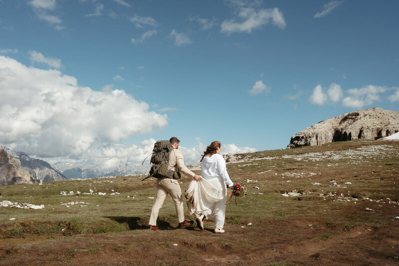 Couple doing a Wedding hike in the Dolomites, groom holds brides dress while they hike
