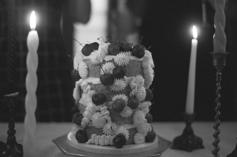 Photo is black and white - a tall cake sits on a stand with lit candles in the background. The cake is naked style with the layers completely exposed, and is decorated with fresh cherries and dollops and squiggles of icing.