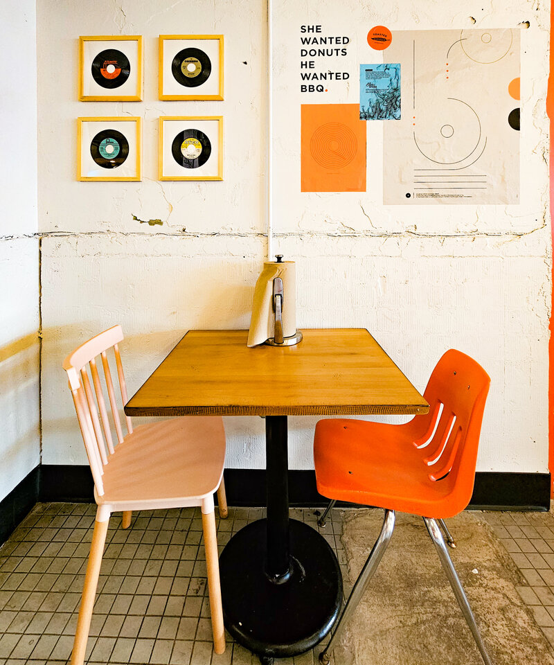 two mismatched chairs at a brown wood table at smoke and donuts in orlando florida with a background wall of vinyl recors and magazine clippings