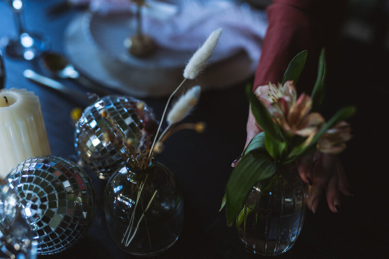 A hand arranging a vase of flowers on a table with disco balls, candles, and cutlery at a luxury event planning setup in Northwest Arkansas.