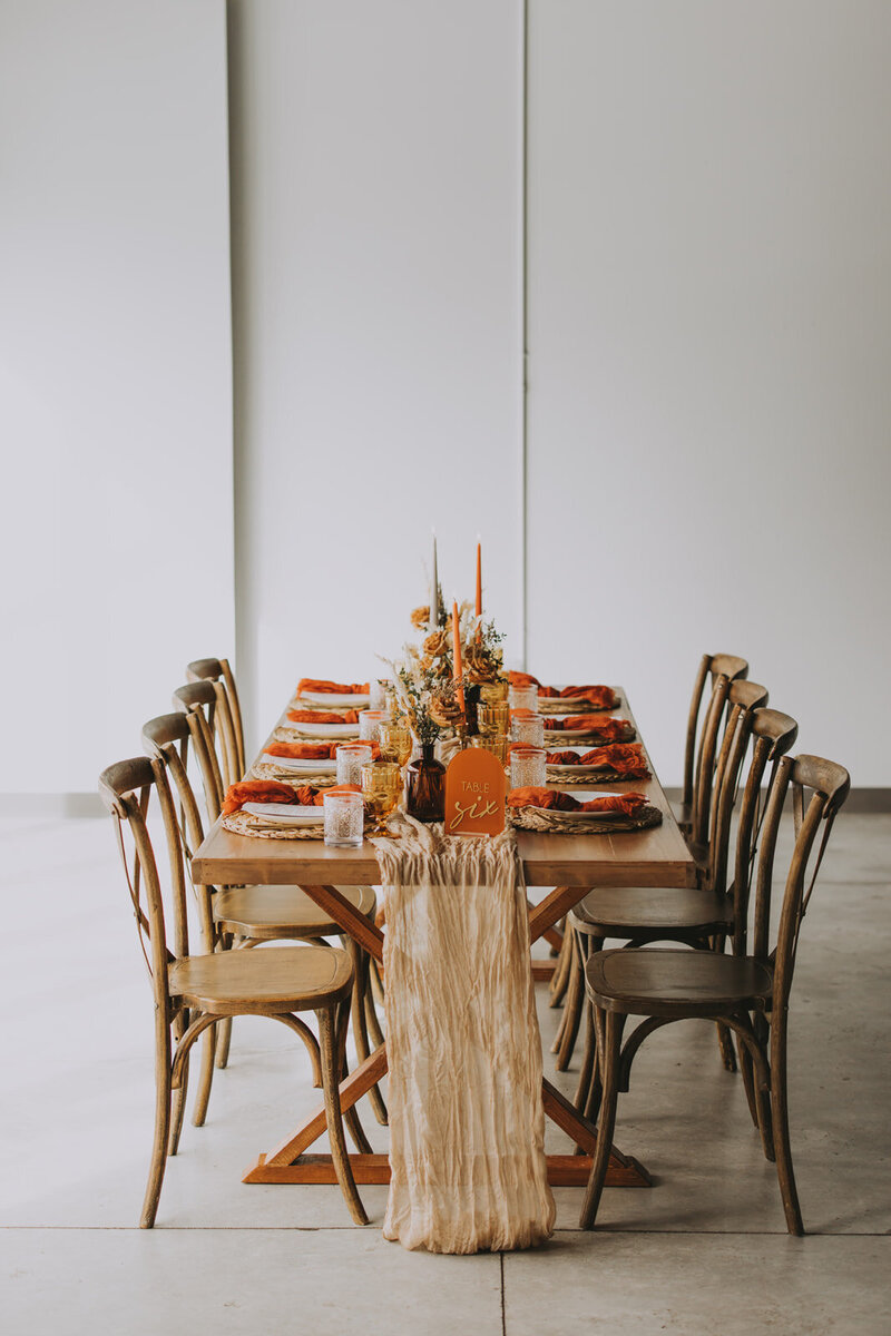 Rustic wooden wedding table styled with burnt orange napkins, amber glassware, neutral gauze table runner, and dried floral centerpiece for a modern earthy tones for a  reception