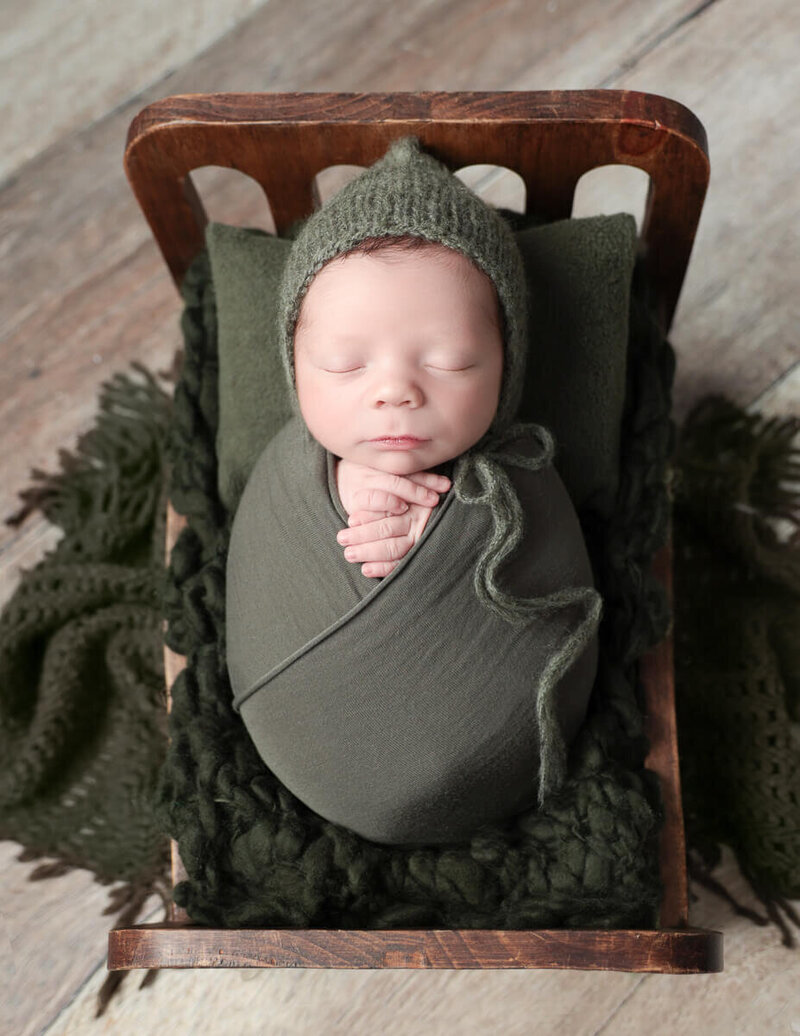 Newborn boy peacefully sleeping on a wooden bed, adorned with a soft green bonnet, in a cozy photography studio in Rochester, NY.