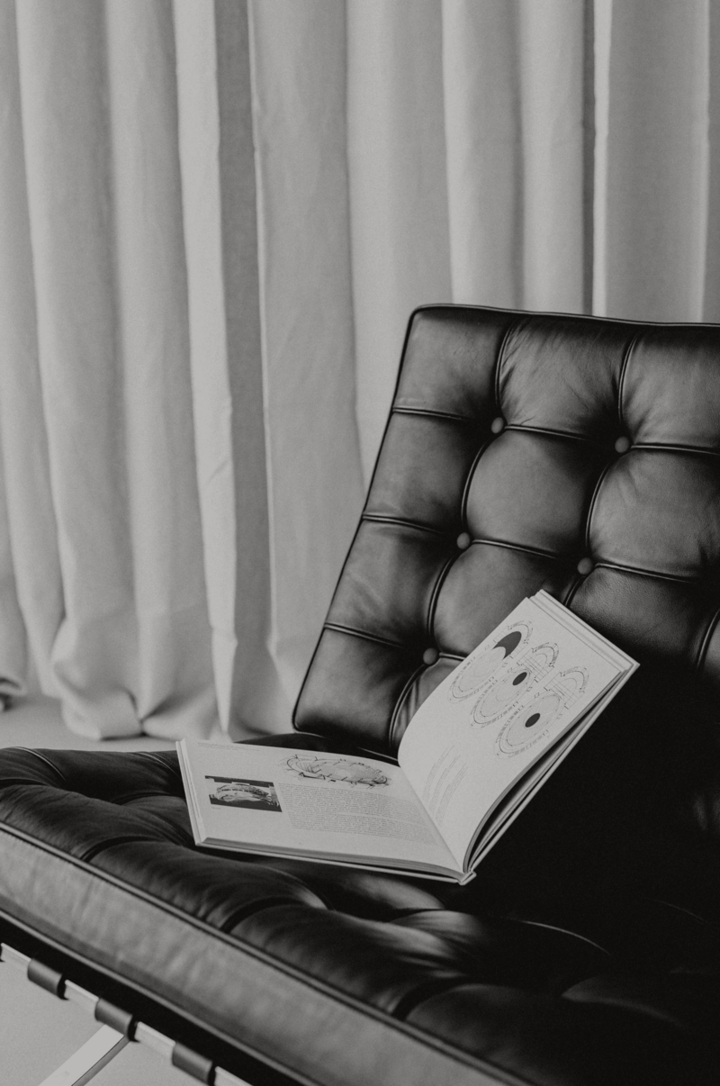 Egy fekete-fehér felvétel egy elegáns, bőr fotelről, melyen egy nyitott könyv fekszik. A minimalista stílusú enteriőrben egy ablak természetes fényt enged a helyiségbe.