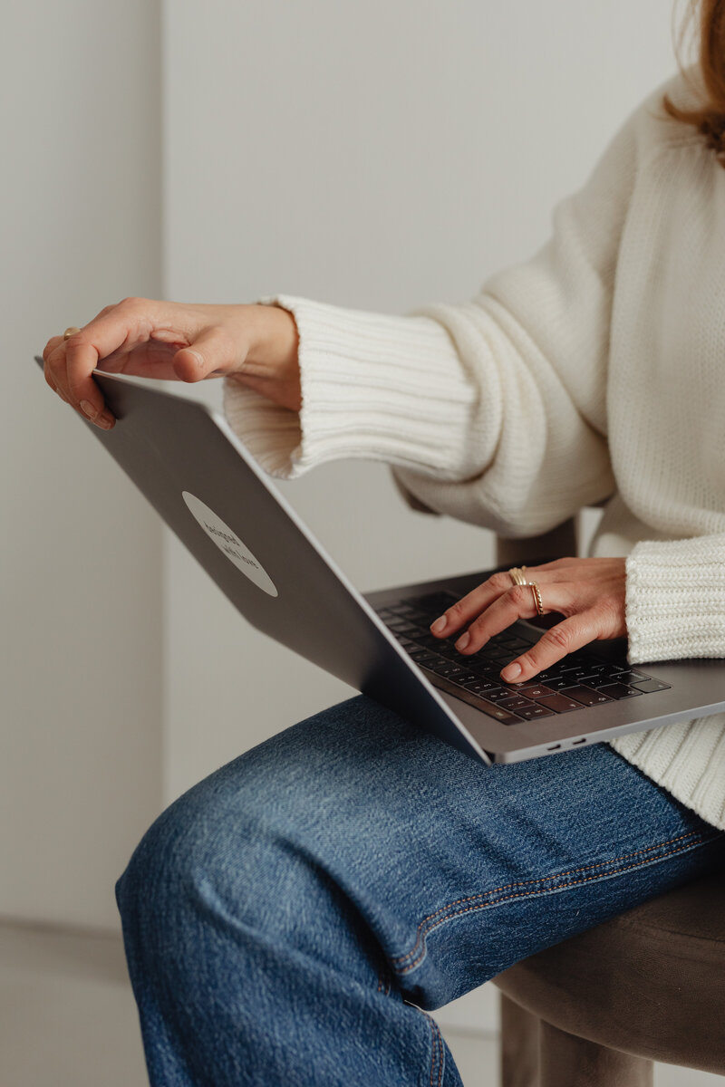woman in white sweater and jeans typing on laptop