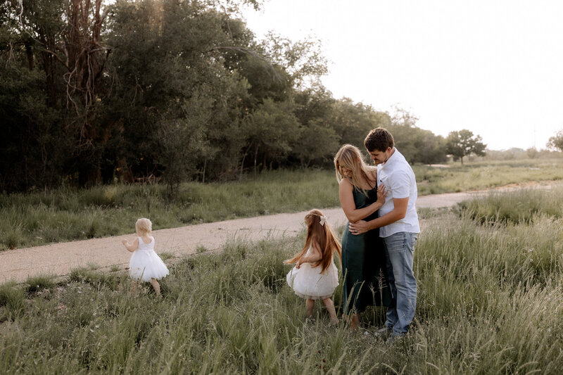 family of 4 photo taken in Denver City texas at the park dad has his arms wrapped around waist with girls running around them