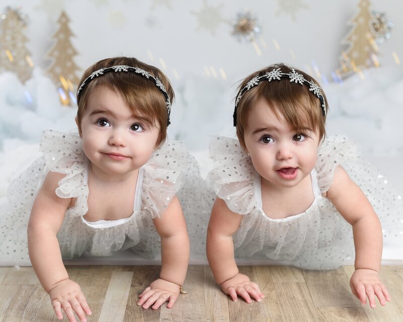 Twin baby girls dressed in sparkling white outfits with snowflake headbands, crawling on a wooden floor surrounded by a winter wonderland theme with gold trees and snowflakes.