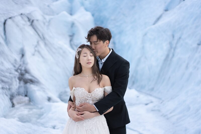Closeup of a couple in wedding attire on a glacier in Iceland with blue ice behind them. The groom is embracing the bride and they both have closed eyes.
