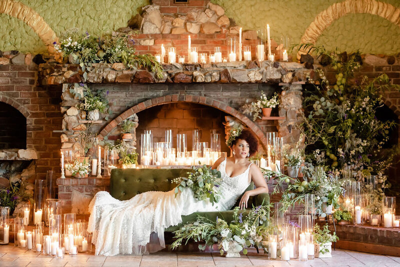 Stunning bride in a lace gown lounges on a green velvet loveseat in front of a glowing candlelit stone fireplace with lush floral decor at grbic events in st. louis