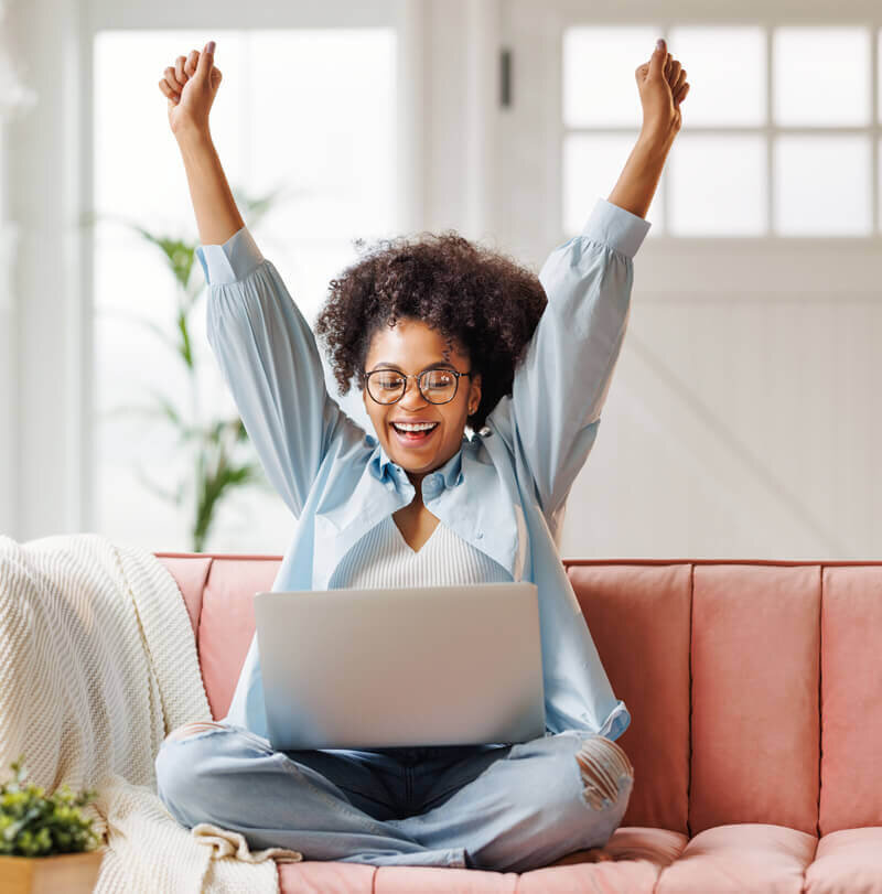 Business woman sitting cross-legged on a sofa with her arms raised over her head and smiling at her laptop