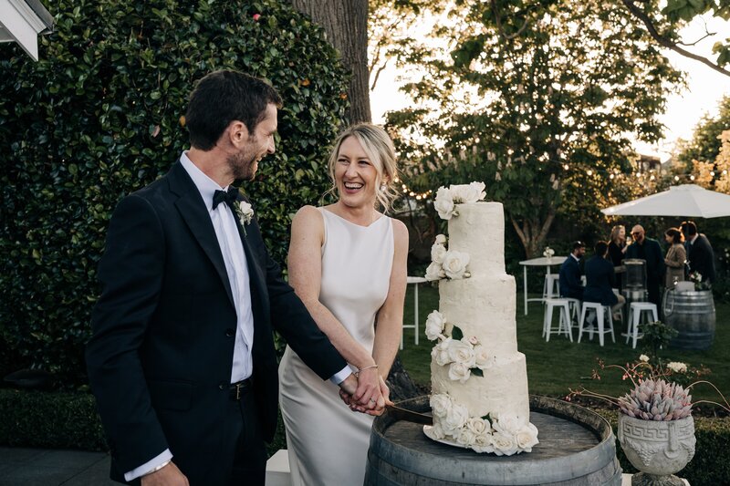 bride and groom cut three tier ivory wedding cake with white roses in backyard wedding