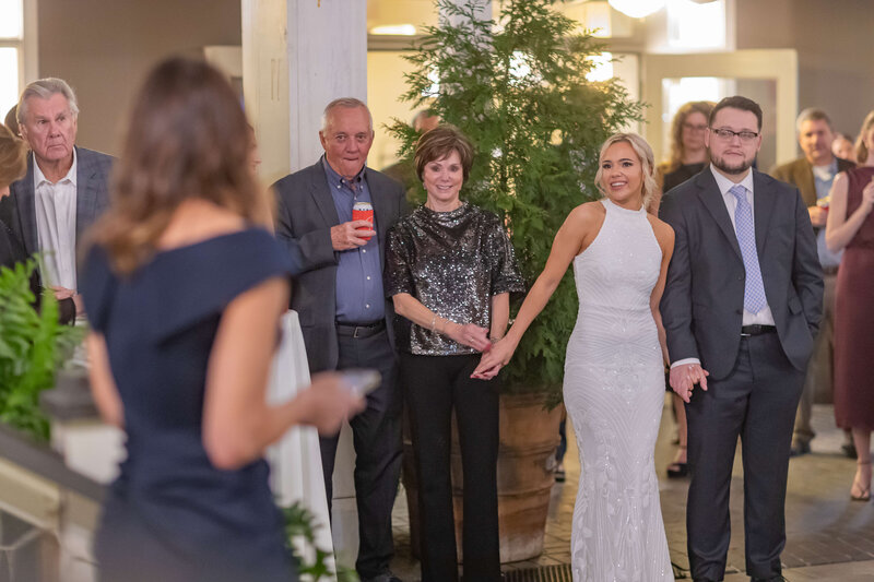 A bride in a white gown smiles while holding hands with a groom in a suit. They stand with two older adults, listening to a woman giving a speech at a formal outdoor event. Other guests are visible in the background.