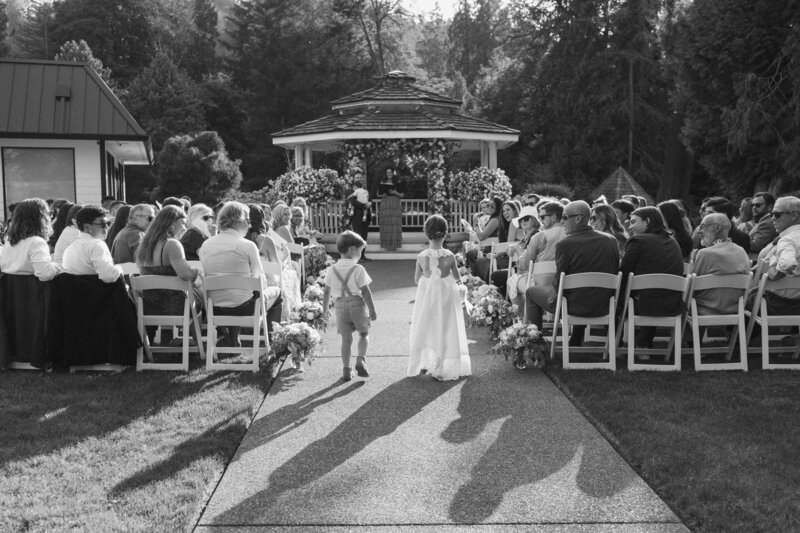 Two children walk toward a flower-covered gazebo at an outdoor ceremony, a joyful scene in candid Seattle wedding photography.