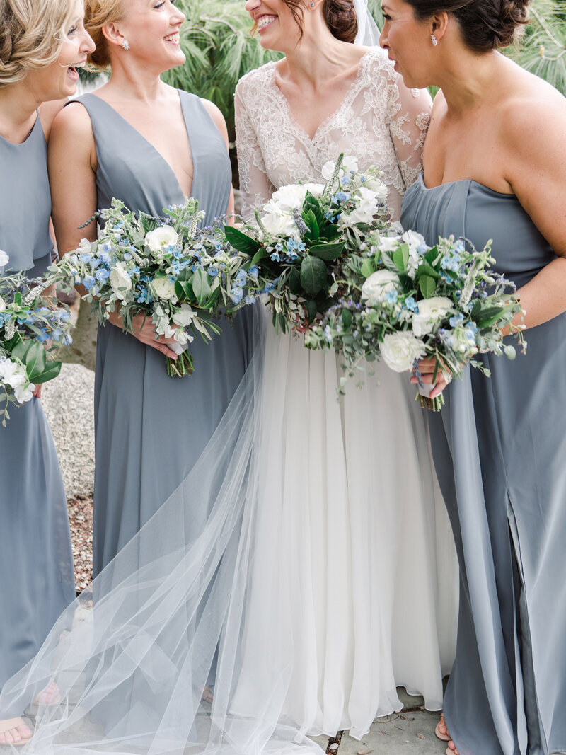 Bride in lace gown with bridesmaids in gray dresses holding white blue and green spring bouquets of tweedia and peonies