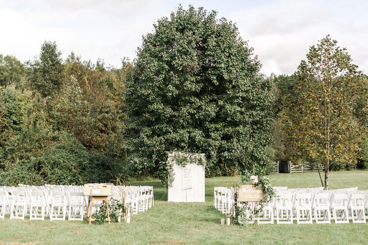 outdoor boho whimsical ceremony in open field under a large tree with white chairs and greenery at glasgow farm in fredericksburg, viriginia planned by cait and co events
